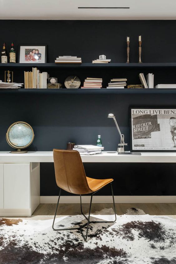 a contemporary home office with a black wall and open shelves, a white desk, an amber leather chair and an animal skin rug