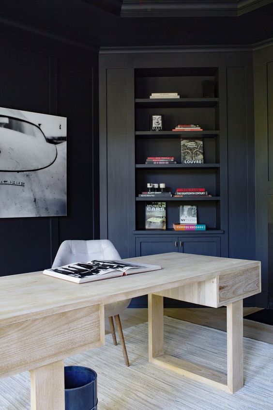 a contrasting home office with navy walls and built-in furniture, a bleached wooden desk and a statement artwork on the wall
