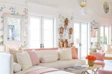 a feminine space with wallpaper accents, a creamy corner sofa, creamy and pink pillows, pink chairs and a floral chandelier