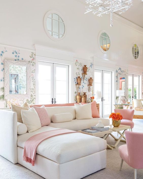 a feminine space with wallpaper accents, a creamy corner sofa, creamy and pink pillows, pink chairs and a floral chandelier