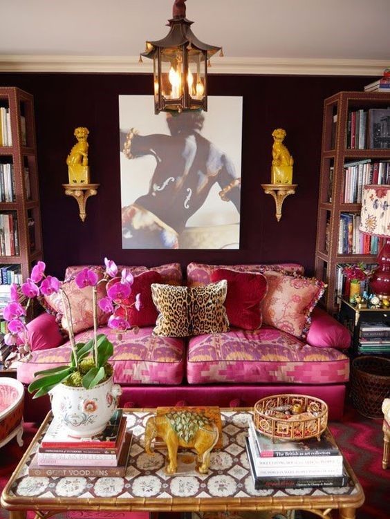 a maximalist and feminine living room with black walls, a pink and gold sofa with pillows, an inlaid coffee table, unique art and lamps