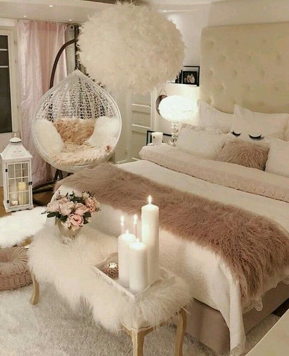 a neutral and blush feminine bedroom with a white upholstered bed, a furry bench, a suspended chair and a feather chandelier