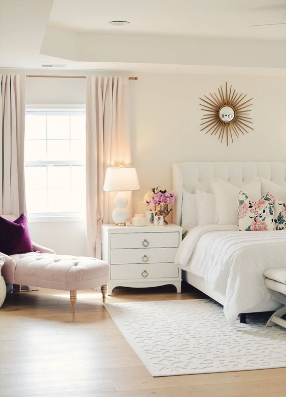 a neutral feminine bedroom with a white upholstered bed, white nightstands, a blush couch and a purple pillow and blush curtains