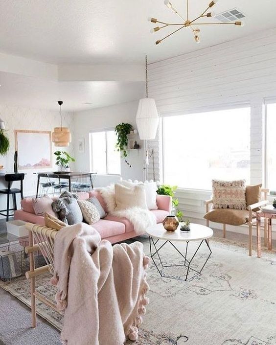 a pretty Scandi living room with a feminine feel, a pink sofa with lots of pillows, a table with a geo base, a woven chair and other chairs, potted greenery
