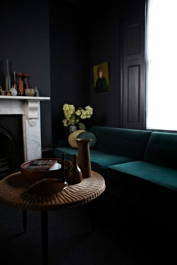 a refined dark masculine room with dark walls, a forest green sofa, a fireplace and a wicker coffee table