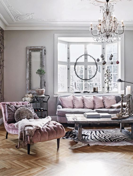 a refined feminine living room with a grey sofa with lilac pillows a mauve lounger a crystal chandelier a rough wood coffee table and greenery