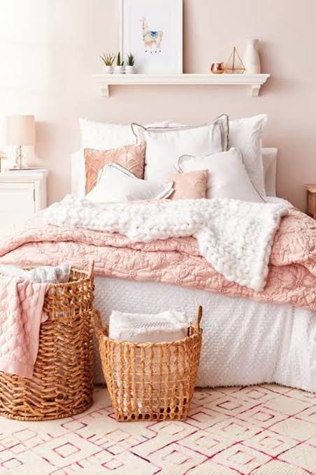 a relaxed feminine bedroom with blush walls, simple furniture, white and pink bedding, a shelf with art and a couple of baskets