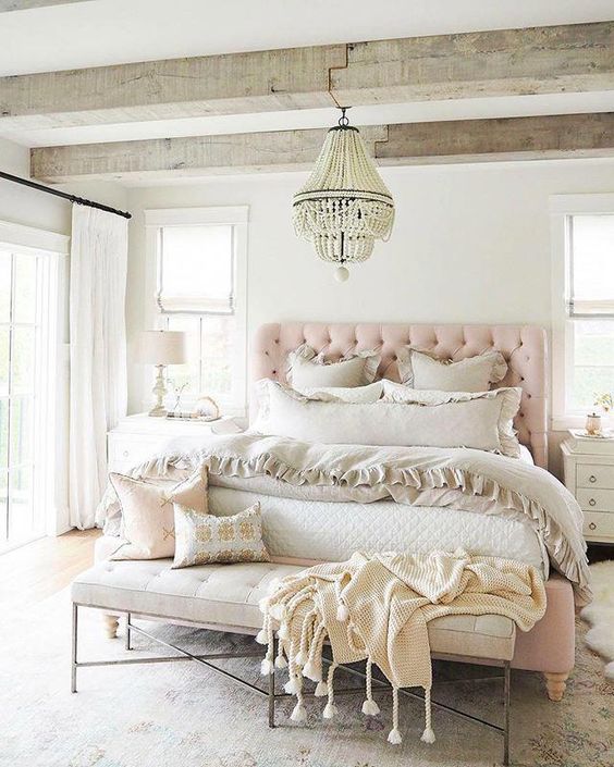 a relaxed feminine bedroom with wooden beams, a pink upholstered bed, a leather bench, neutral bedding and a chic chandelier