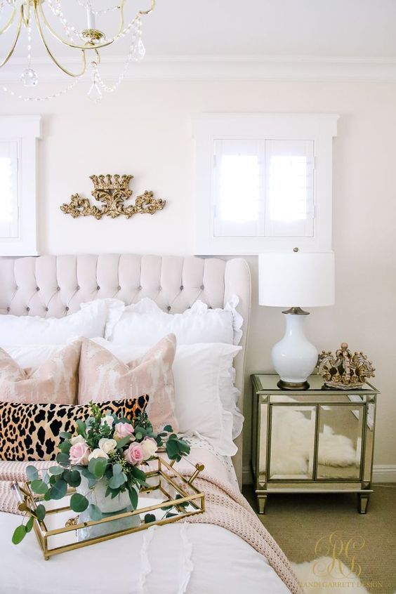 a romantic and feminine bedroom with a neutral upholstered bed, blush and white bedding, a crystal chandelier and a mirror nightstand