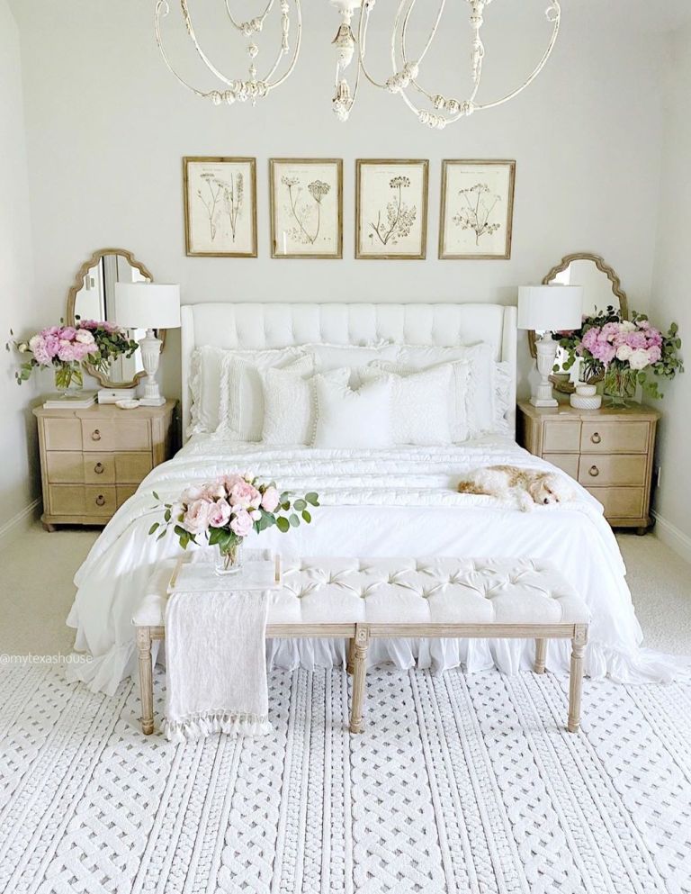 a shabby chic feminine bedroom with a white bed and bench, botanical artworks, wooden nightstands, a chic chandelier and blooms