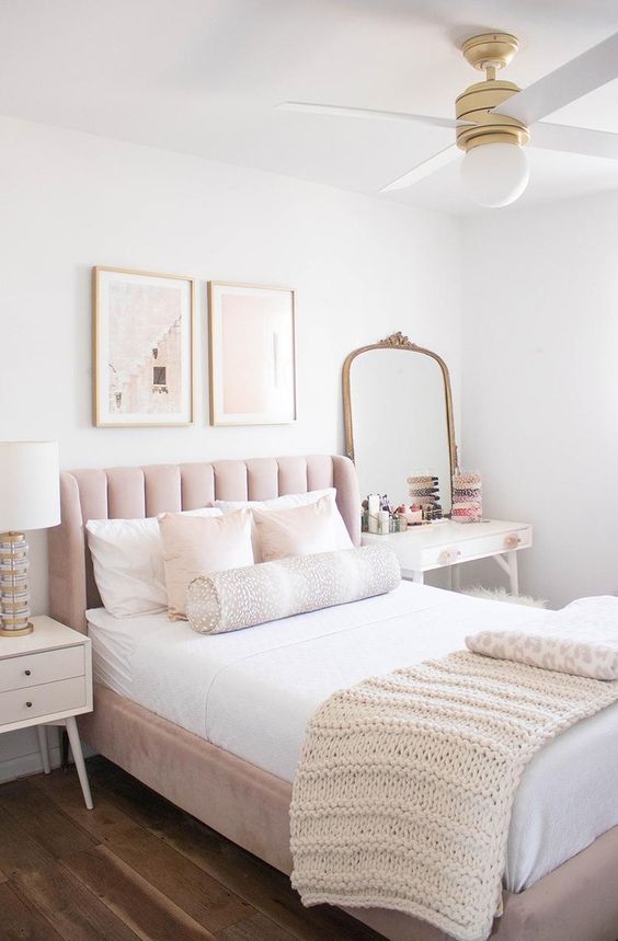 a simple feminine bedroom with a blush bed, white nightstands, a mirror in a brass frame and cute artworks