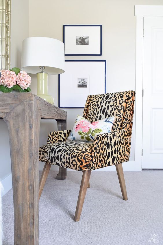 a small working space with a wooden desk, a leopard print chair, a floral pillow, a gallery wall and a table lamp