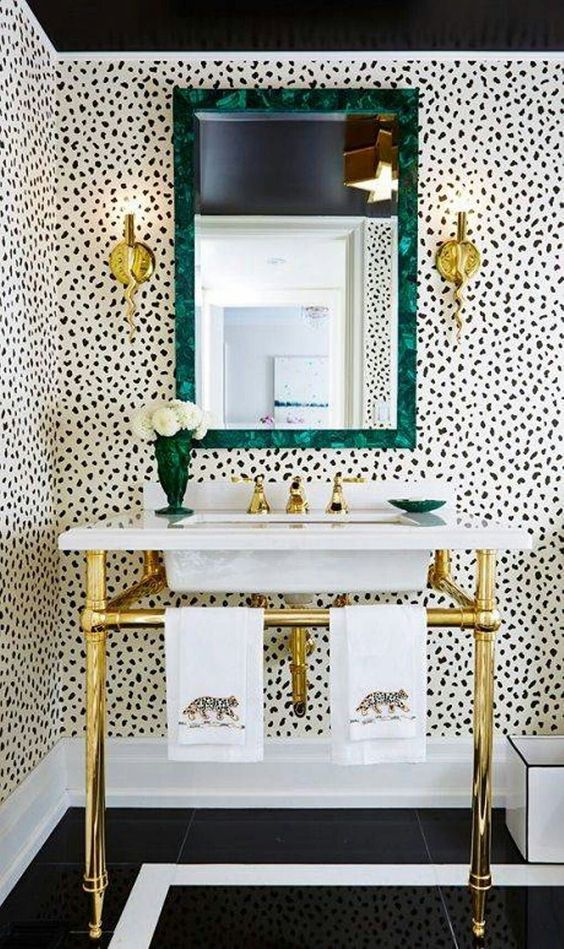 a stylish bathroom with Dolmatin print walls, a free standing sink on gold legs, a mirror in a green frame and gold sconces is wow
