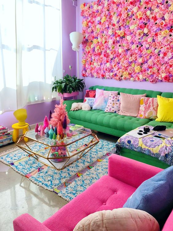 a super colorful maximalist living room with a bright faux flower wall, a green sofa, a hot pink chair, colorful pillows, a diamond-shaped glass table