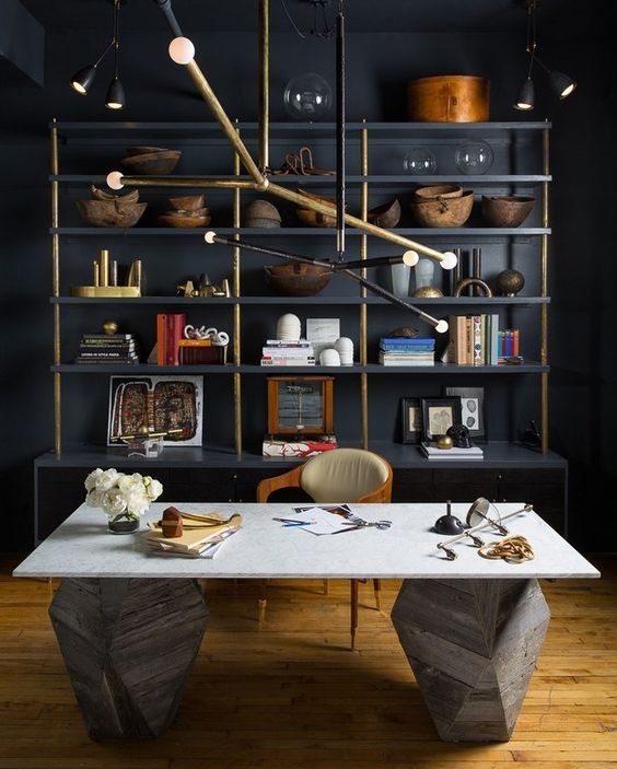 a unique home office with blakc walls, an open shelving unit, a sculptural stone desk and a unique metal chandelier