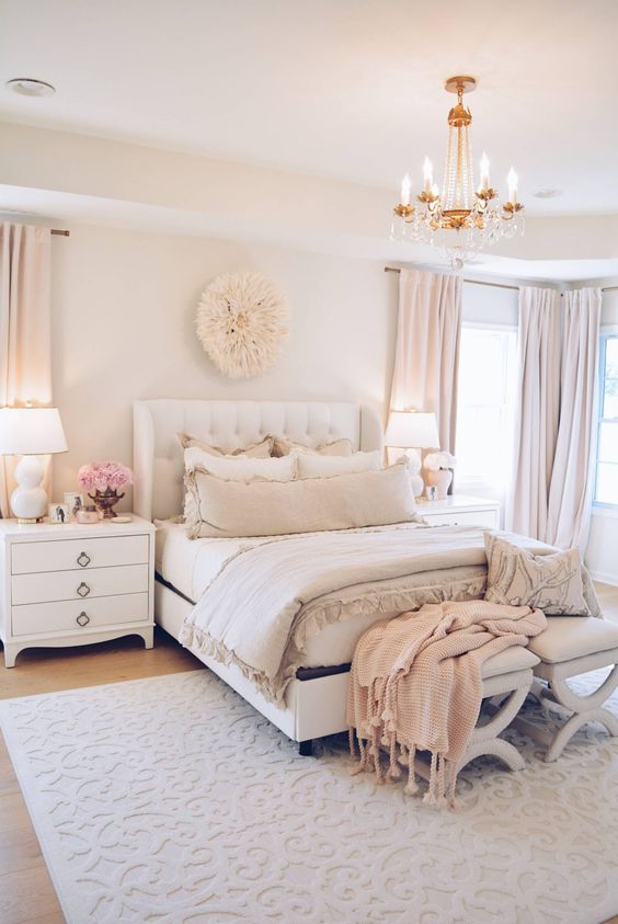 a welcoming feminine bedroom in neutrals and blush, with an upholstered bed, neutral furniture, a crystal chandelier and touches of blush