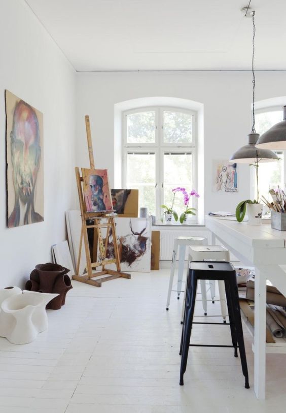 a Nordic art studio with a large white table, a row of stools, an easel, some bright artwork, potted plants and blooms