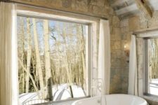 a barn bathroom clad with stone, a panoramic window, an antler chandelier, a free-standing tub and a bright rug