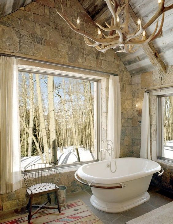 a barn bathroom clad with stone, a panoramic window, an antler chandelier, a free-standing tub and a bright rug