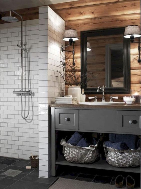 a barn bathroom with a wooden wall, a vintage grey vanity, a white tile clad shower space and a mirror in a black frame