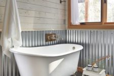 a barn bathroom with whitewashed wooden planks, corrugated steel on the walls, a clawfoot tub, a large window and a fluffy rug