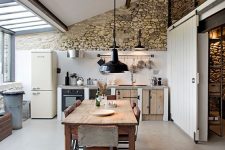 a barn kitchen with a stone accent wall, a glazed one, planked cabinets, dark wooden beams and a vintage dining set of wood and metal