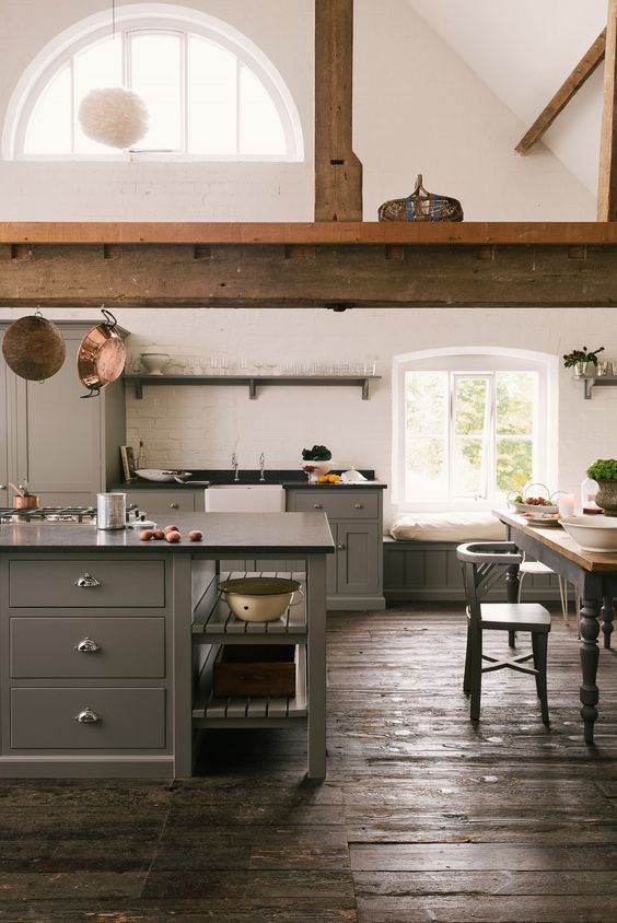 a barn kitchen with grey cabinets, a large wooden beam, black countertops, a vintage dining set and white brick walls