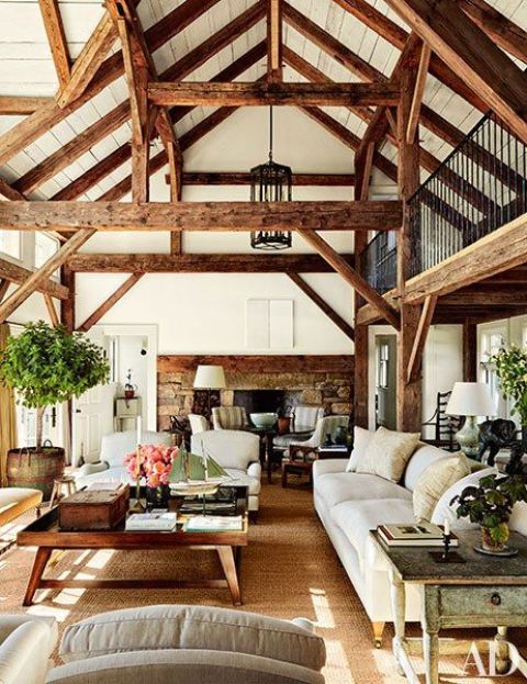a barn living room with multiple wooden beams, a fireplace clad with stone, white seating furniture, a stained coffee table and potted greenery