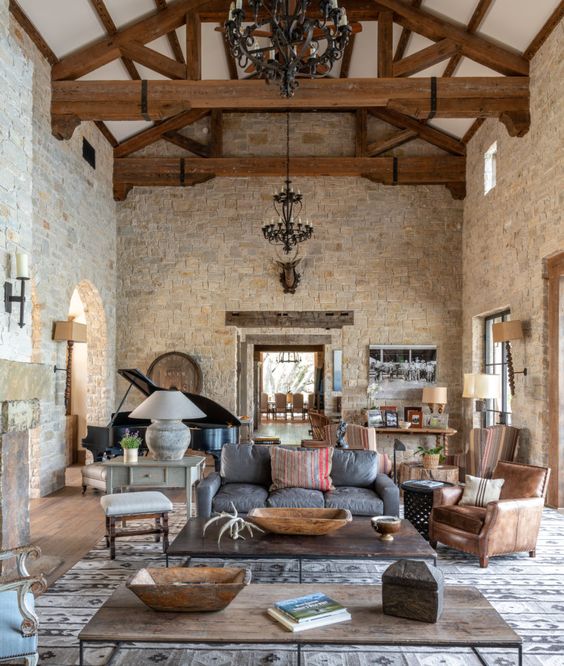 a barn living room with stone walls, wooden beams on the ceiling, brown, grey and white furniture, a piano, refined metal chandeliers and low coffee tables