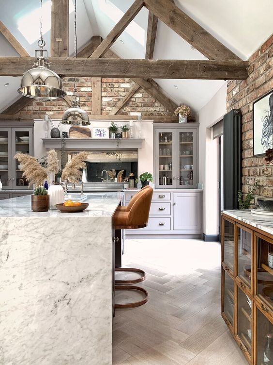 a beautiful barn kitchen with reclaimed wooden beams, metal pendant lamps, white cabinets, a stone kitchen island and chic stools