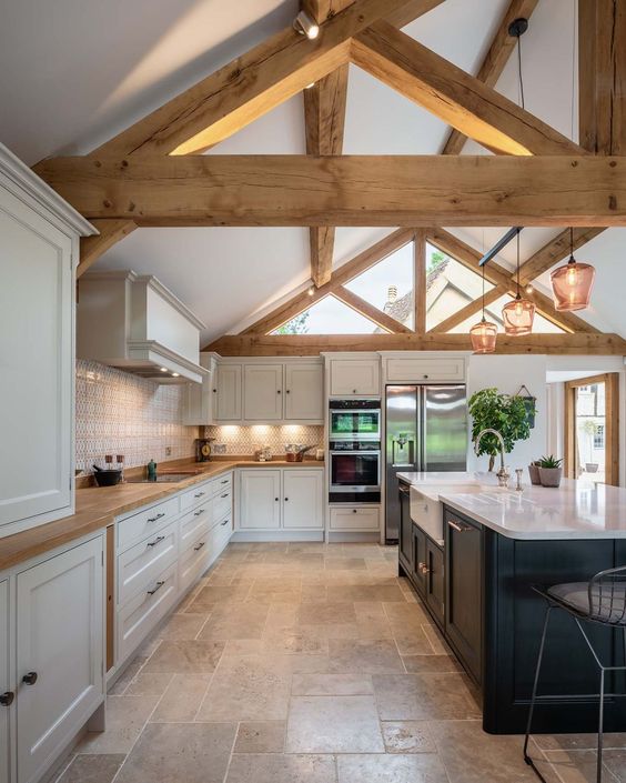 a beautiful barn kitchen with wooden beams, white shaker style cabinets, a black kitchen island and elegant glass pendant lamps
