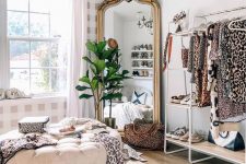 a bold feminine closet with an open storage unit, a shoe shelf, a mirror in an ornated frame, a neutral tufted pouf and a potted plant
