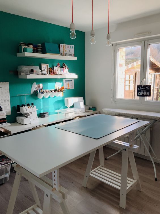 a bright artist studio with a turquoise accent wall, a trestle desk and a white table, pendant bulbs and some decor