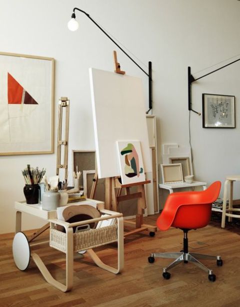 a catchy and bold home artist studio with a wooden easel and a stylish cart with supplies, a red chair, a comsole table and some art