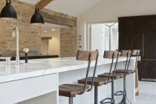 a contemporary barn kitchen with light-stained wooden beams, a white planked kitchen island, vintage industrial stools and black pendant lamps