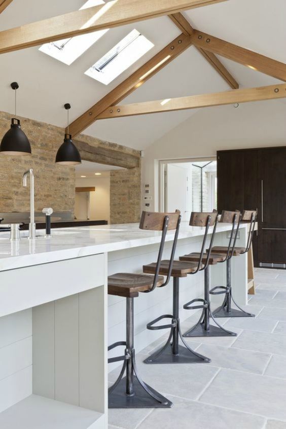 a contemporary barn kitchen with light-stained wooden beams, a white planked kitchen island, vintage industrial stools and black pendant lamps