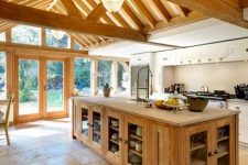a cozy barn kitchen with wooden beams on the ceiling, white cabinets, a light-stained kitchen island, a crystal chandelier and a tile floor