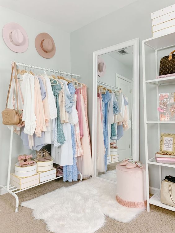 a cute pastel closet with light blue walls, an open makeshift closet, a large mirror, layered rugs, a pink stool and hats as decor