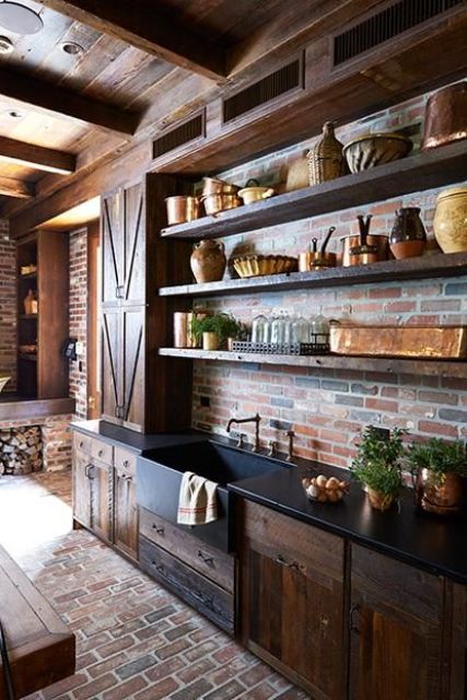a dark kitchen with red brick walls and floor, dark reclaimed wood cabinets and matching shelves plus potted greenery