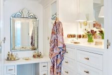 a delicate and chic small walk-in closet with white closed storage units and drawers, a white vnaity, a couple of mirrors and touches of gold