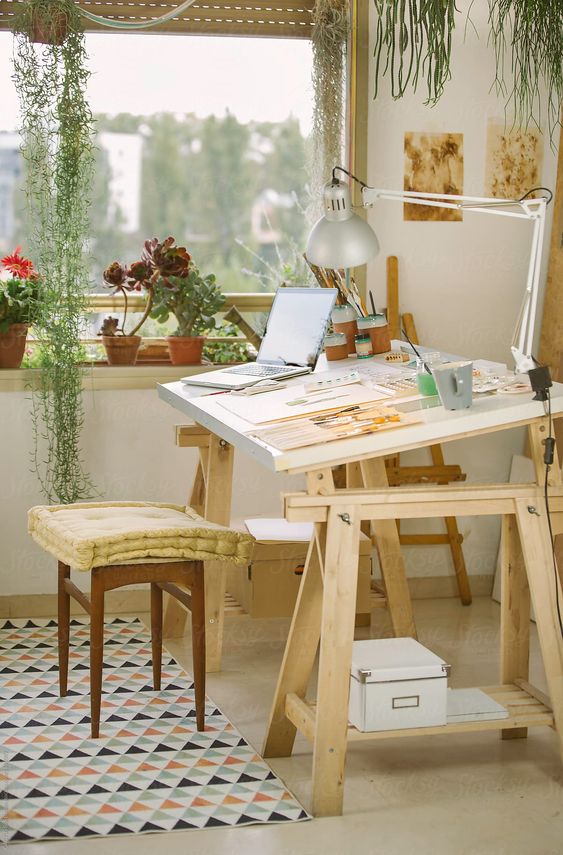 a home artist studio with potted plants and succulents, a large stained desk, a stool, an easel and some artwork