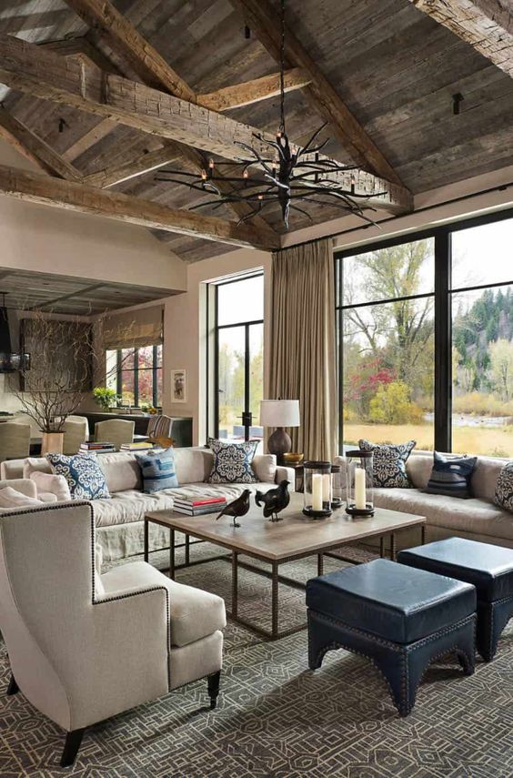 a lovely barn living room with a reclaimed wooden ceiling and beams, neutral seating furniture, blue poufs, an antler chandelier and amazing landscapes
