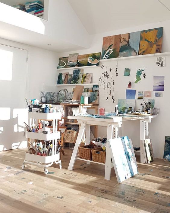 a lovely home art studio with a large table, an IKEA cart with supplies, ledges and a shelf, some bright artwork is adorable