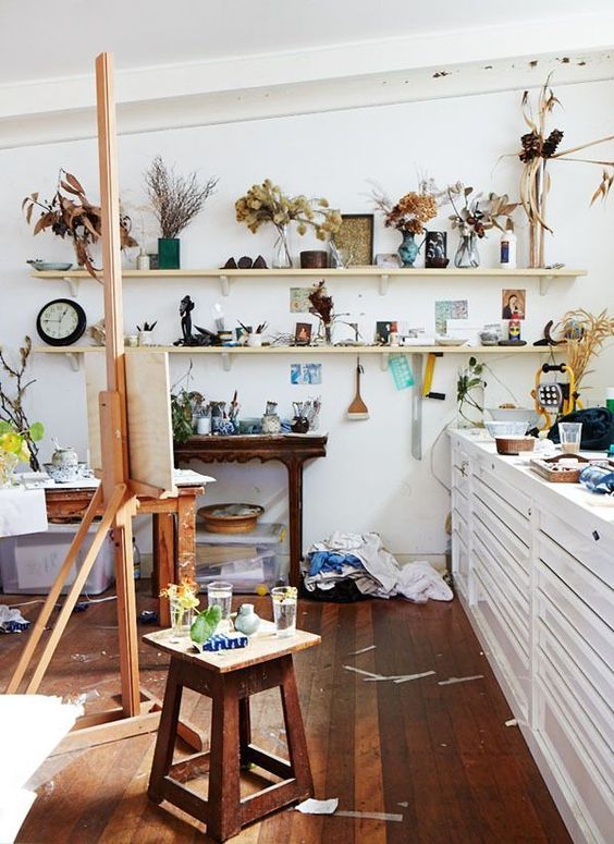 a messy and eclectic home art studio with open shelves with decor and dried foliage, a white storage cabinet, an easel, stools and console tables