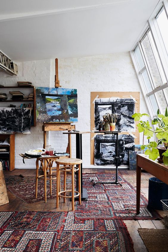 a mid-century modern home artist studio with a glazed wall, colorful boho rugs, an easel, some stools, a large table and artwork