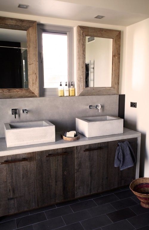 a minimalist barn bathroom with concrete walls, a weathered wood vanity, concrete sinks and mirrors in wooden frames