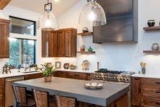 a modern barn kitchen with stained cabinets, white stone and concrete countertops, a shiny hood, wooden beams and pendant lamps