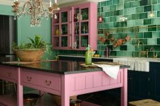 a navy kitchen with hot pink upper cabinets and a matching kitchen island, black stone countertops, a bold emerald tile backsplash and a vintage crystal chandelier