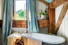 a pretty barn bathroom with wooden plank walls, a floor and ceiling, wooden beams, a blue clawfoot tub and blue curtains on the window