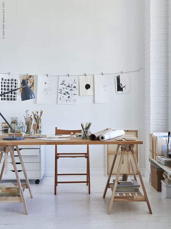 a pretty light-filled studio with a trestle desk, a chair, a file cabinet, a table lamp, some shelves and lots of artwork